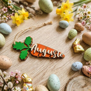Étiquette de Pâques en forme de carotte personnalisée en contreplaqué de haute densité, entourée d'œufs colorés, de fleurs de printemps et de petits lapins dorés sur un fond en bois clair.