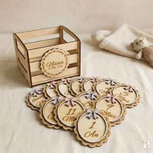 Un coffret en bois personnalisé pour bébé, de style petite cagette, avec une étiquette gravée au nom de "Naïna" et la date "06 Mai 2026". Devant le coffret vide, un ensemble de cartes étapes en bois de forme ovale avec des bordures festonnées style dentelle est disposé en éventail sur un tissu en lin clair. Chaque carte est ornée d'un petit nœud en relief de couleur rose ou lilas et indique un mois ou une étape, comme "Bienvenue", "11 Mois", "1 An", "9 Mois", "5 Mois". En arrière-plan, un lange en tissu et un petit ours en peluche tricoté complètent la scène.